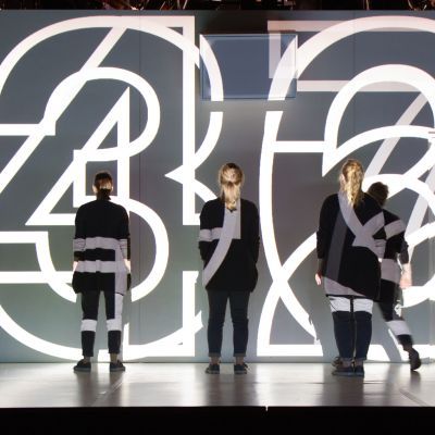 Large white neon numbers projected to the back of a stage over performers standing facing the back